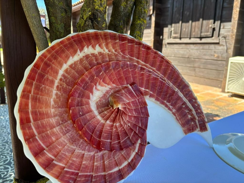 Lonchas de jamón ibérico en espiral presentadas sobre una copa blanca junto a un tronco, en el exterior