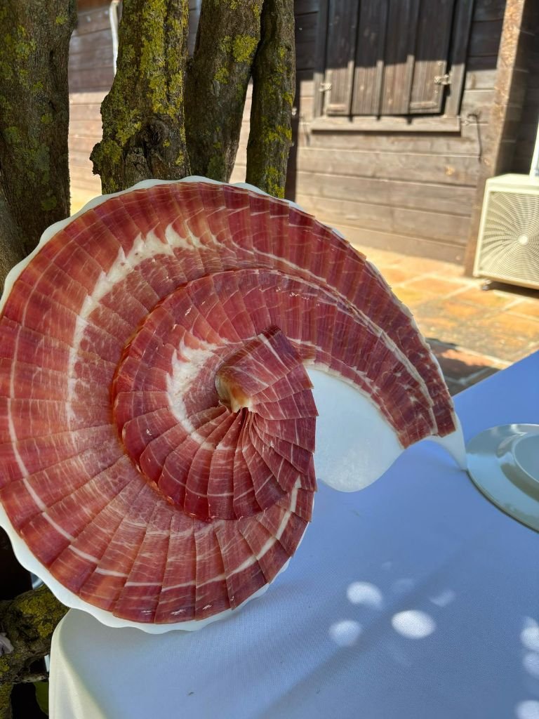 Plato blanco con lonchas de jamón en espiral sobre copa, apoyado en una mesa con mantel azul, al aire libre